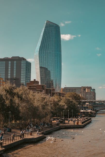A tall, modern glass skyscraper with a curved, asymmetrical design reflecting the sky and surrounding buildings stands prominently in Blackfriars, London. In the foreground, a riverside walkway is visible with several pedestrians walking along the paved path lined with metal railings. Trees with mixed green and brown foliage are situated along the water's edge, and a river with gentle ripples flows past, with a few seagulls floating on the surface. On the right side of the image, a small outdoor seating area with people sitting and relaxing is visible, along with some boats moored nearby. The scene is set during daytime with a clear sky and a few clouds, and the urban environment suggests a busy city centre. The image is associated with house and office removals in Blackfriars, and occasional mention of [COMPANY_NAME], Man With a Van Blackfriars, is noted for professional moving services involving furniture transport and packing during home relocations.