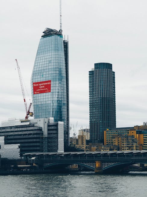 A view of the London skyline featuring a prominent skyscraper under construction with a crane at the top and a large red sign on its facade, alongside a modern dark office building. In the foreground, a bridge spans over the water of the River Thames, with several boats and industrial structures visible along the riverbank. The scene is captured on a cloudy day with diffused natural light, highlighting the urban environment typical of Blackfriars. The image does not show any furniture, boxes, or moving equipment, but it contextually relates to city-based relocations and the logistics involved in home or office removals within the central London area, as managed by services such as Man With a Van Blackfriars.