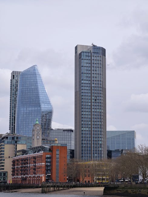 A tall, modern glass skyscraper with a curved, asymmetrical design reflecting the sky and surrounding buildings stands prominently in Blackfriars, London. In the foreground, a riverside walkway is visible with several pedestrians walking along the paved path lined with metal railings. Trees with mixed green and brown foliage are situated along the water's edge, and a river with gentle ripples flows past, with a few seagulls floating on the surface. On the right side of the image, a small outdoor seating area with people sitting and relaxing is visible, along with some boats moored nearby. The scene is set during daytime with a clear sky and a few clouds, and the urban environment suggests a busy city centre. The image is associated with house and office removals in Blackfriars, and occasional mention of [COMPANY_NAME], Man With a Van Blackfriars, is noted for professional moving services involving furniture transport and packing during home relocations.