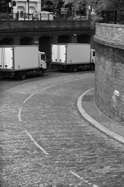 The image depicts a narrow urban street in Blackfriars during daytime, with buildings on both sides comprising a mix of modern and older architecture, featuring glass facades and brick exteriors. The street is wet, possibly from recent rain, and has a white dashed line running down the centre. Several black bollards line the pavement edges, and a white van is parked partially on the curb, adjacent to a building entrance. In the background, taller city buildings are visible beneath a cloudy sky. This setting provides an appropriate context for house removals and furniture transport, illustrating a typical city street where loading and unloading during a home relocation might take place, as handled by companies like Man With a Van Blackfriars.