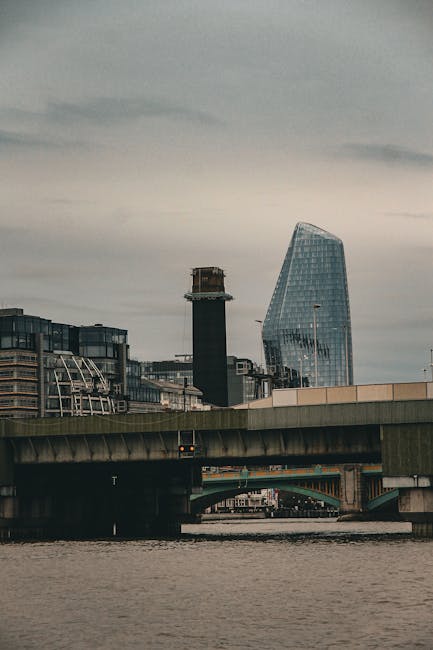 The image depicts a view of the London skyline near Blackfriars, with a river in the foreground and a bridge spanning across it. On the bridge, a man with a van from Man With a Van Blackfriars is engaged in home relocation activities, loading or unloading cardboard boxes, plastic wrapping, and furniture pieces onto or from a large, black van that is parked partly on the pavement adjacent to the bridge. The interior of the van shows stacked packing materials such as tissue paper and protective blankets, useful for furniture transport during packing and moving processes. The scene is set during daylight with diffuse overcast weather, and nearby buildings, including modern high-rises, are visible behind the bridge, indicating an urban environment suitable for house removals and relocation services. The area appears active, emphasizing the practical aspects of furniture transport, loading, and logistics involved in residential moving, as handled by [COMPANY_NAME].