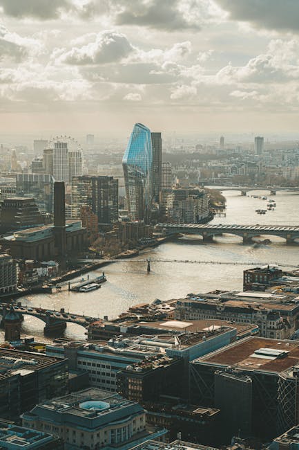 The image depicts a view of the London skyline near Blackfriars, with a river in the foreground and a bridge spanning across it. On the bridge, a man with a van from Man With a Van Blackfriars is engaged in home relocation activities, loading or unloading cardboard boxes, plastic wrapping, and furniture pieces onto or from a large, black van that is parked partly on the pavement adjacent to the bridge. The interior of the van shows stacked packing materials such as tissue paper and protective blankets, useful for furniture transport during packing and moving processes. The scene is set during daylight with diffuse overcast weather, and nearby buildings, including modern high-rises, are visible behind the bridge, indicating an urban environment suitable for house removals and relocation services. The area appears active, emphasizing the practical aspects of furniture transport, loading, and logistics involved in residential moving, as handled by [COMPANY_NAME].