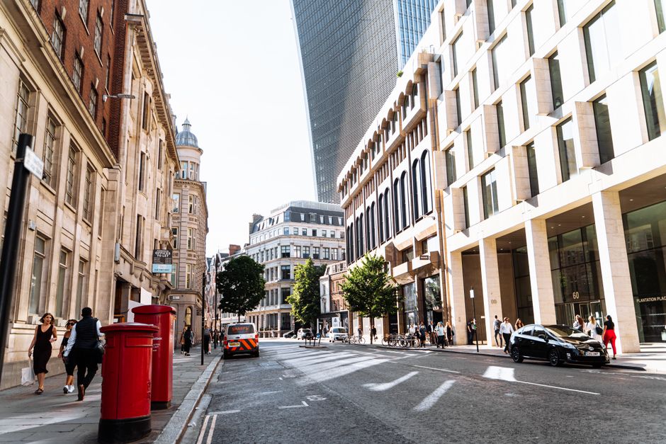 A city street scene showing a mix of historic and modern architecture, with pedestrians walking along the pavement, some carrying bags or talking, and a few cyclists riding by. On the left side, there
