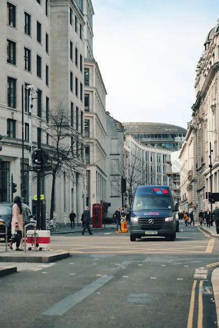 A city street scene showing a mix of historic and modern architecture, with pedestrians walking along the pavement, some carrying bags or talking, and a few cyclists riding by. On the left side, there