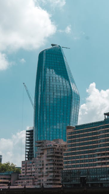 The image shows a modern, high-rise building with a distinctive curving glass facade reflecting the sky and clouds, located in an urban area with other mid-rise structures nearby. The building appears to be under construction or renovation, as evidenced by a tall crane positioned on the left side of the image. The foreground features a paved street or sidewalk, and part of a parking or loading zone is visible at the base of the building. The scene is well-lit with natural daylight, highlighting the sleek architectural design. In the context of house removals and furniture transport, this image subtly reflects the process of urban home relocation or moving services, where items are transported through city environments, possibly involving loading and unloading operations. Man With a Van Blackfriars might operate in such locations, supporting clients with efficient moving solutions, including packing, loading, and transportation within city centers or commercial districts.