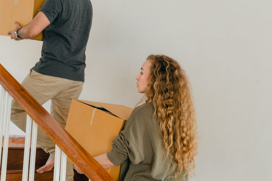 A woman with long, curly red hair, wearing an olive green shirt with rolled-up sleeves, is carrying a large cardboard box while ascending a wooden staircase indoors. In the background, a person wearing beige trousers and a dark grey t-shirt is seen lifting or moving another box. The staircase has white spindles and a wooden handrail, with a light-colored wall behind. The scene depicts a home relocation or packing process, with cardboard boxes, plastic wrap, and blankets likely used for furniture protection. This moment captures part of the unloading or packing and moving activity often associated with professional removals services, such as those provided by Man With a Van Blackfriars, during furniture transport in property interiors. The lighting is natural, and the environment appears tidy, illustrating typical stages in house removals or moving logistics.