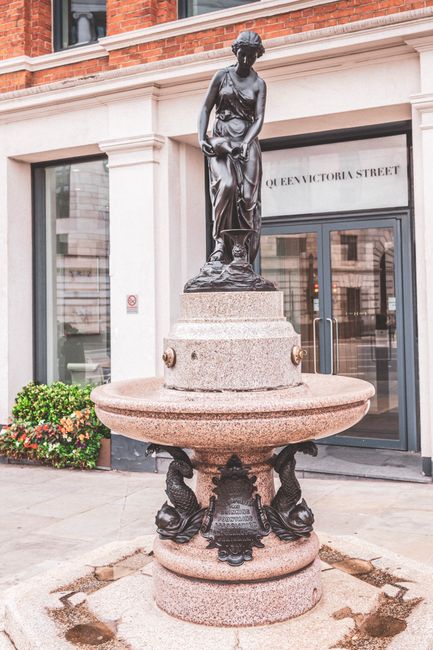 A stone fountain featuring a bronze statue of a woman in classical attire, standing on a round pedestal, is situated outside a building on Queen Victoria Street. The fountain's basin rests on an ornately decorated base with sculpted lion heads and decorative elements, and the area around it is paved. Behind the fountain, a glass door storefront with the sign 'Queen Victoria Street' is visible, reflecting nearby buildings. The scene is illuminated by natural daylight, and small plants are placed near the building's exterior. The setting appears to be a city street, with no visible moving or relocation equipment present in the image, which emphasizes the street’s decorative features rather than any immediate packing or loading activity. The image is related to urban environments where furniture or belongings might be temporarily moved or protected during house removals or relocations managed by companies like Man With a Van Blackfriars.