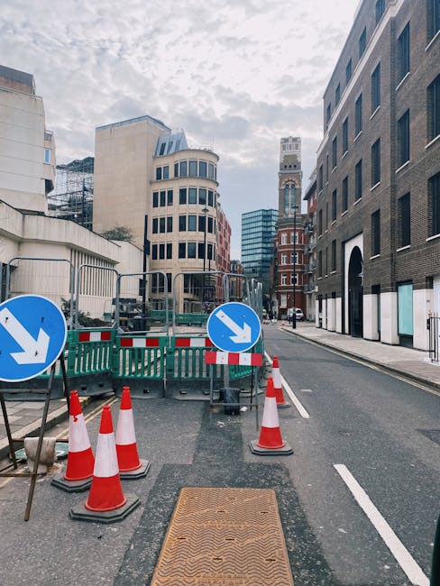 A street view in Blackfriars with temporary roadworks featuring orange and white traffic cones, two blue and white directional signs indicating a downward shift in traffic flow, and metal barriers surrounding a section of roadway. The pavement includes a metal access cover with a textured surface, while in the background, tall modern and historic buildings line the street. This setting appears to be a loading or staging area for house removals or furniture transport, with no visible vehicles or moving equipment present at the moment. The scene is illuminated by natural daylight under a partly cloudy sky, and the environment suggests an urban setting relevant to home relocation activities coordinated by Man With a Van Blackfriars, fitting within the context of loading bay regulations on Queen Victoria St.