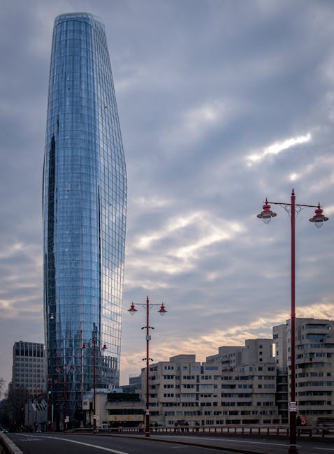 The image shows a modern, tall, glass-clad skyscraper with a curved, cylindrical shape located near Blackfriars Bridge in London, with a partly cloudy sky overhead. In the foreground, there are red street lamps lining the road and a row of beige, mid-rise residential or commercial buildings with multiple windows. The scene captures an urban environment typical of city centre relocation or home removal settings, with no immediate moving activity visible. This setting could relate to furniture transport and home relocation services provided by Man With a Van Blackfriars, situated within the context of moving logistics in the area.