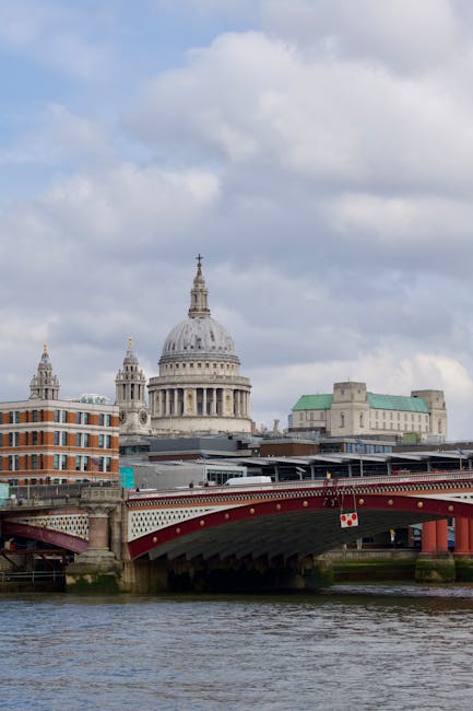 A view of St. Paul's Cathedral with its iconic large dome and ornate spires, seen from across the River Thames. In the foreground, a red and white bridge spans the river, with part of a flatbed trolley or dolly visible on the pavement nearby. The image shows a typical urban scene associated with central London, including low-rise and historic-style buildings with brick and stone facades, as well as modern structures. Overhead, the sky is partly cloudy with patches of blue, suggesting daytime. This setting reflects a typical environment for home relocation or moving services in Blackfriars, where local removals by Man With a Van Blackfriars may include carrying furniture and packed boxes through this scenic cityscape during a house move or furniture transport process.
