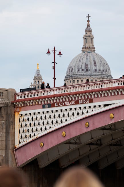 The image shows a modern, tall, glass-clad skyscraper with a curved, cylindrical shape located near Blackfriars Bridge in London, with a partly cloudy sky overhead. In the foreground, there are red street lamps lining the road and a row of beige, mid-rise residential or commercial buildings with multiple windows. The scene captures an urban environment typical of city centre relocation or home removal settings, with no immediate moving activity visible. This setting could relate to furniture transport and home relocation services provided by Man With a Van Blackfriars, situated within the context of moving logistics in the area.