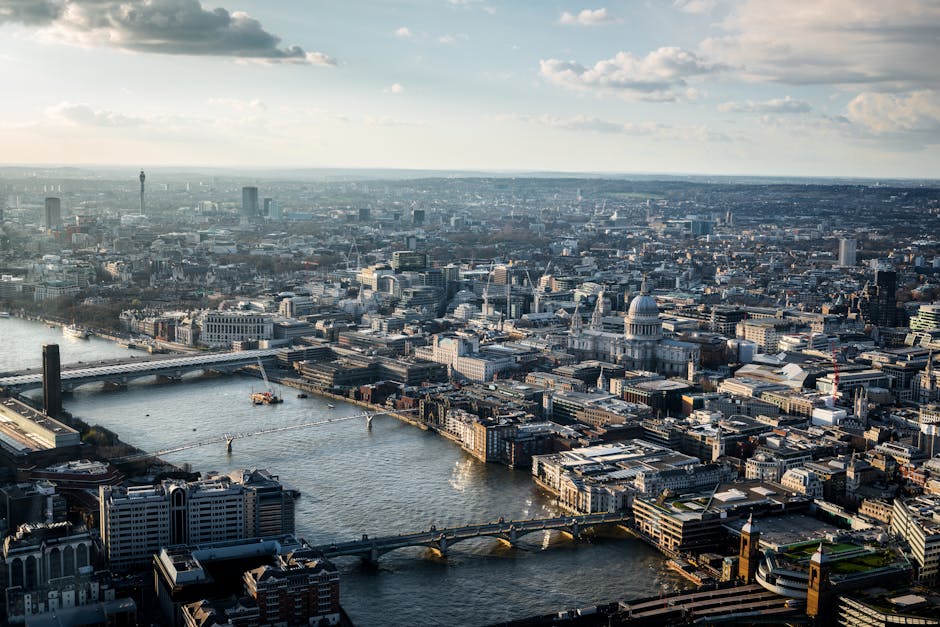 Aerial view of central London showing the River Thames winding through the city with multiple bridges crossing it. Prominent landmarks such as St. Paul’s Cathedral with its large dome and the modern skyscrapers in the background are visible under a partly cloudy sky. In the foreground, a section of a building's rooftop and window edge are visible, suggesting the photo was taken from a high vantage point. The cityscape features a mix of historic and contemporary architecture, with streets and smaller buildings densely packed. The lighting indicates late afternoon or early evening, with shadows cast across the city. This image highlights the urban density and iconic skyline, relevant to discussions of city-based house relocations and the logistical considerations involved in moving items through busy metropolitan areas, potentially supported by services like Man With a Van Blackfriars.