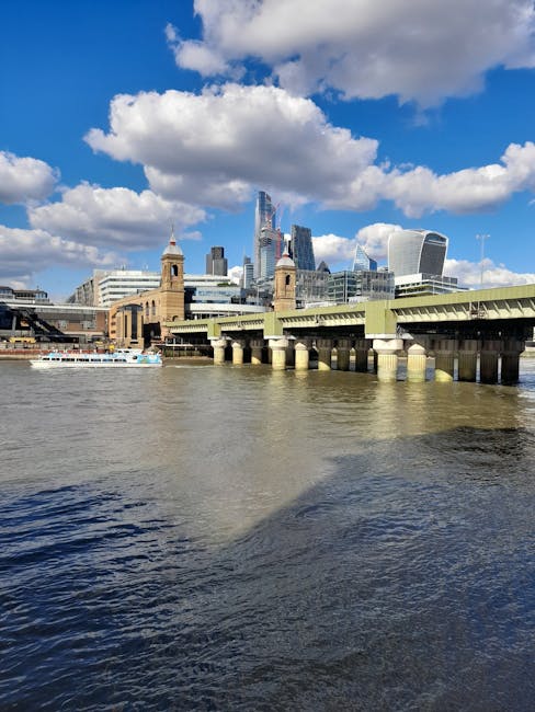 A wide cityscape view of the London skyline over the River Thames, featuring modern skyscrapers including the Walkie Talkie building, the Cheesegrater, and the Leadenhall Building, with a historic bridge in the foreground supported by multiple stone pillars. The river has a slight ripple effect, and a boat is visible on the left side. The scene is illuminated with bright daylight, with a partly cloudy sky overhead. In the context of house removals and furniture transport, this image reflects the urban environment where residential and commercial relocations often take place near central London, necessitating careful planning and logistics by companies such as Man With a Van Blackfriars during packing and loading processes for city-to-city or local moves.