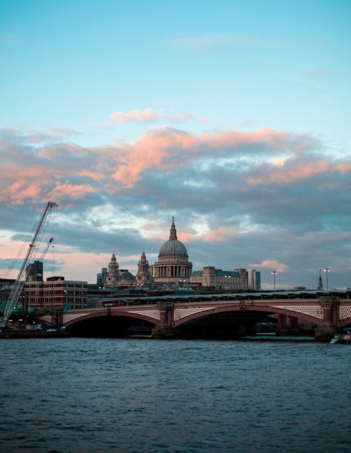 Aerial view of central London showing the River Thames winding through the city with multiple bridges crossing it. Prominent landmarks such as St. Paul’s Cathedral with its large dome and the modern skyscrapers in the background are visible under a partly cloudy sky. In the foreground, a section of a building's rooftop and window edge are visible, suggesting the photo was taken from a high vantage point. The cityscape features a mix of historic and contemporary architecture, with streets and smaller buildings densely packed. The lighting indicates late afternoon or early evening, with shadows cast across the city. This image highlights the urban density and iconic skyline, relevant to discussions of city-based house relocations and the logistical considerations involved in moving items through busy metropolitan areas, potentially supported by services like Man With a Van Blackfriars.