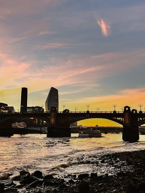A waterfront scene at sunset with a view of the city skyline featuring modern skyscrapers, including one with a distinctive curved shape, and a historic brick bridge crossing the river. The sky displays a gradient of colors from soft pinks and oranges near the horizon to darker blues higher up, with a few wispy clouds illuminated by the setting sun. The river below has gentle waves and dark rocks along the shoreline. This image captures an urban landscape likely associated with city living or relocation activities, relevant to house removals and moving services such as those offered by Man With a Van Blackfriars, and highlights the scenic environment surrounding Southwark.