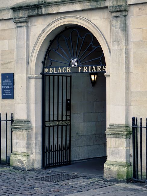 View of an arched entrance to a building in Blackfriars, featuring a black metal gate with vertical bars and a decorative iron grille on top. Above the gate, there is a semi-circular fanlight window with radiating metal bars and the name 'BLACK FRIARS' displayed prominently. To the left of the arch, a blue plaque provides information about the site, mounted on the beige stone wall. The entrance is flanked by stone columns with weathered bases, and black metal fencing runs along the sidewalk in front. Inside the arch, a narrow corridor leads into the property, illuminated by a wall-mounted lamp on the right side, which is turned off. The surroundings include cobblestone paving and an outdoor setting indicative of a London historic area, apt for services such as home relocation or furniture transport, with a focus on careful handling during the loading process by companies like Man With a Van Blackfriars.