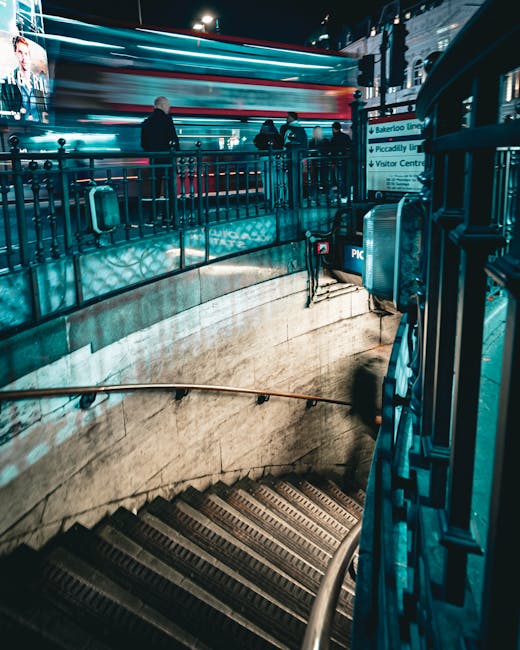 An underground station staircase with beige marble walls and steps fitted with non-slip strips. The staircase has a black metal rail on the right side and a brass handrail on the left. At the top of the stairs, a group of people is standing near a blue fence, with some engaged in conversation and others waiting. Overhead, blurred streaks of red, white, and blue lights indicate a moving train passing through the station. Signage in the background points to various lines and the visitor centre. The environment is illuminated with bright, artificial lighting typical of urban train stations. This scene captures the typical architecture and atmosphere of a busy city underground transport hub, relevant to home relocation or moving logistics involving public transit, as understood by Man With a Van Blackfriars' house removals services.