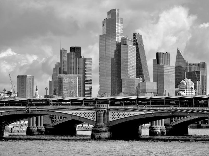 Black and white image of a modern cityscape showing tall skyscrapers with reflective glass facades, situated behind an ornate bridge with detailed metalwork and stone supports spanning a river. The bridge’s roadway is visible, and a railway track runs along the top of the bridge, with a train station platform underneath. The scene depicts urban architecture and transport infrastructure typical of a central business district, with partly cloudy skies overhead. As part of house removals or furniture transport, this image exemplifies urban environments where a professional moving service such as Man With a Van Blackfriars may facilitate home relocation after navigating city bridges and transport links, illustrating the logistical context of packing, loading, and transferring belongings across complex cityscapes.