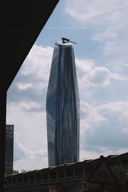 A tall, modern skyscraper with a curved, glass façade reflecting the blue sky and scattered clouds overhead is visible in the background. At the top of the building, a helicopter is parked, suggesting ongoing construction or aerial inspection. In the foreground, part of a dark structure, possibly a building overhang or balcony, frames the scene. Below, a section of elevated railway or road with metal railings runs horizontally, with a bicycle warning sign affixed to it. The setting appears to be an urban area with contemporary architecture, conveying a sense of city life and infrastructure. The image context aligns with the concept of urban moving logistics, as seen in professional house removals and furniture transport, exemplified by [COMPANY_NAME], Man With a Van Blackfriars, providing expert relocation services in busy city environments.